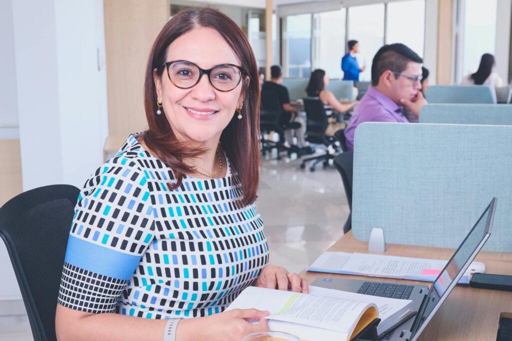 pexels photo 34221316 34221316 Confident woman in a modern office, surrounded by colleagues, working on a laptop.