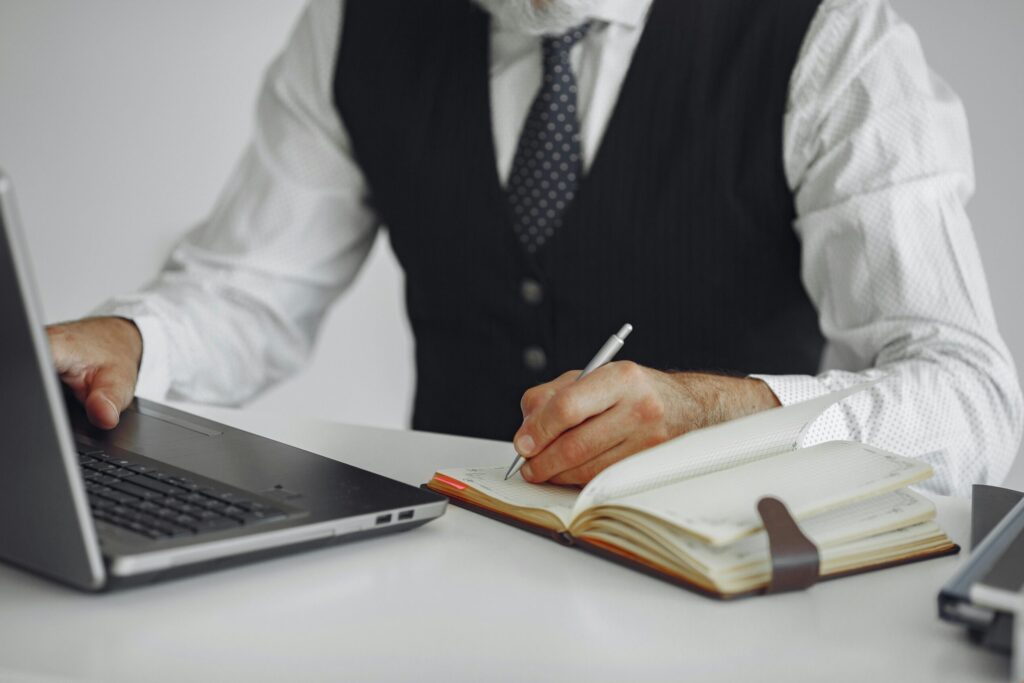 pexels photo 4975658 4975658 Business professional writing notes while working on a laptop at an office desk.