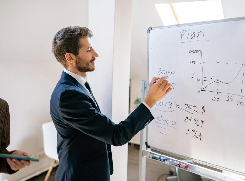pexels photo 7698735 7698735 A businessman in a suit writes financial data on a whiteboard during an office planning session.