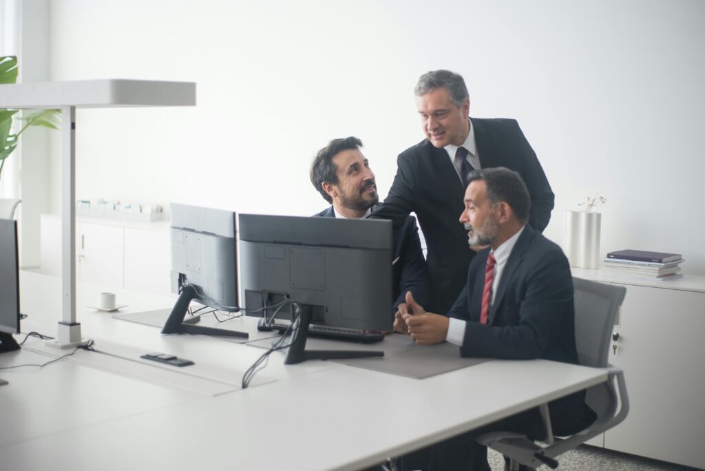 pexels photo 8353822 8353822 Three business executives in suits collaborating around dual monitors in a modern office setting.